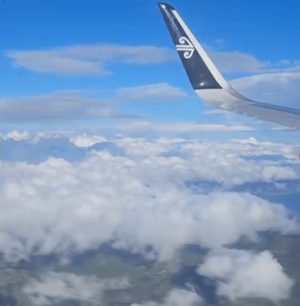 View from an airplane window featuring a blue sky with clouds and part of the airplane wing and tail visible.